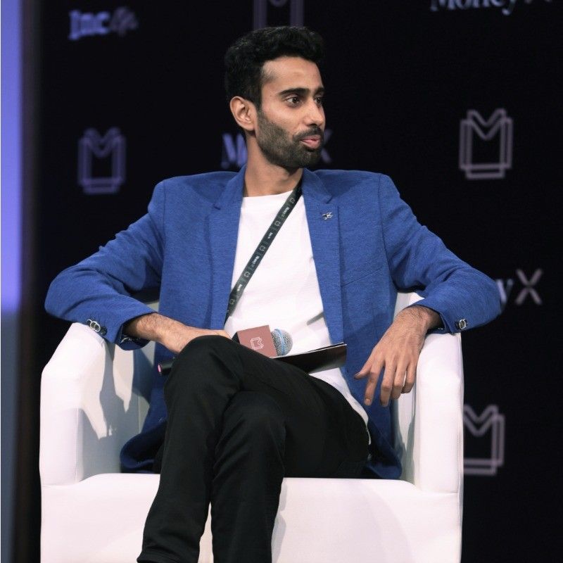 Man in a blue blazer sitting on a white chair on a stage, looking to his left.